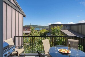 A balcony with a grill, outdoor table, and chairs, overlooking a residential area with houses and a body of water.