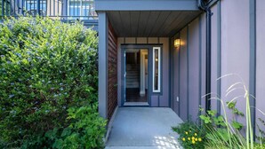 A modern building entrance with a covered walkway, a visible staircase, and a small garden area with various plants. - 3 Bedroom Sooke Townhome (Sooke)