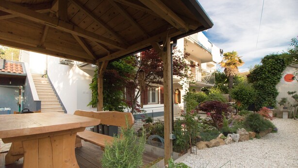 Un'area esterna riparata con tavolo e panche in legno, immersa in un giardino ricco di piante e con un vialetto in ghiaia.