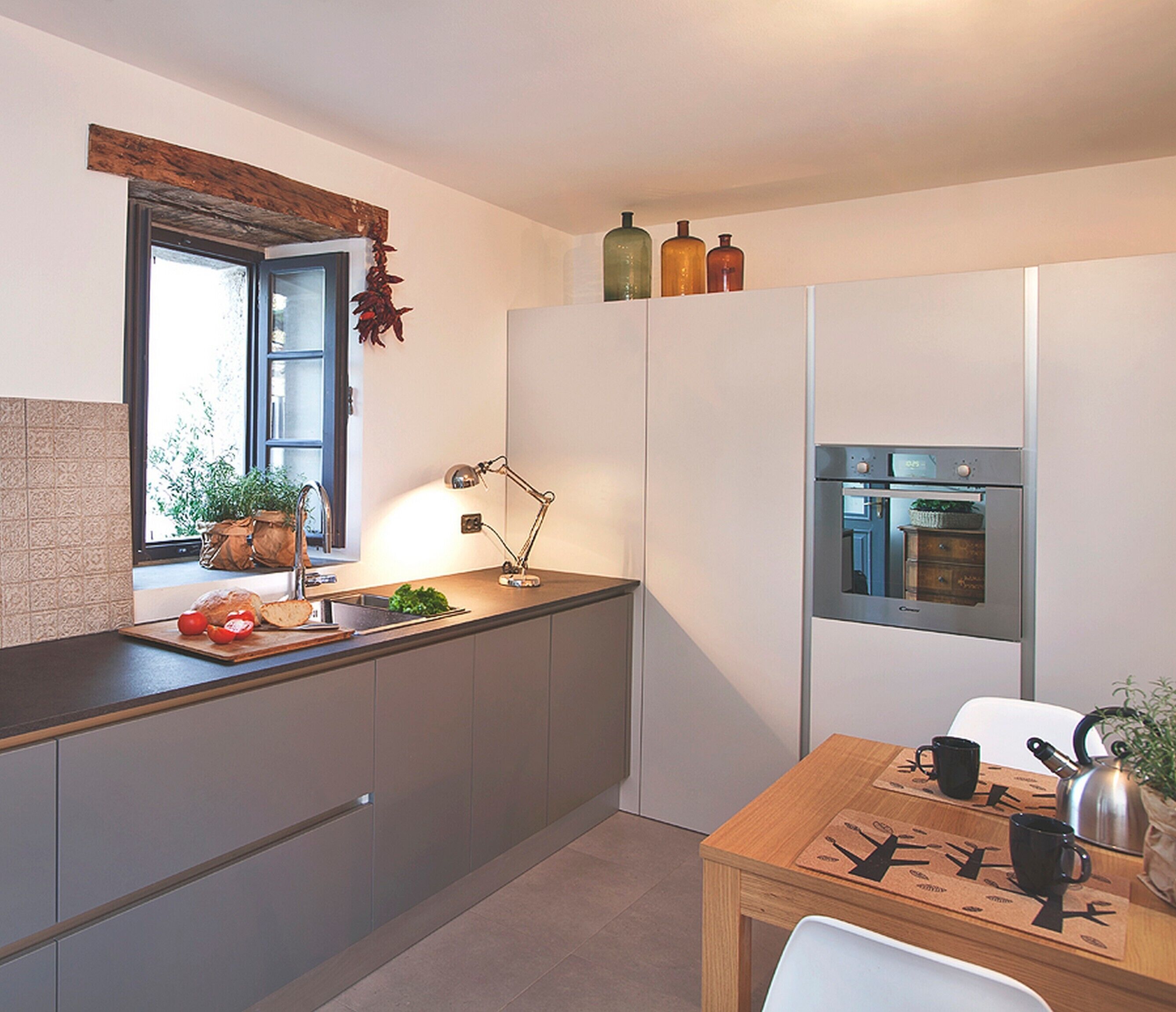 Eine moderne Küche mit grauen Schränken, einer Holzarbeitsplatte, einem Fenster mit Blick, einer Spüle, einem Wasserkocher und einem kleinen Esstisch mit Stühlen.