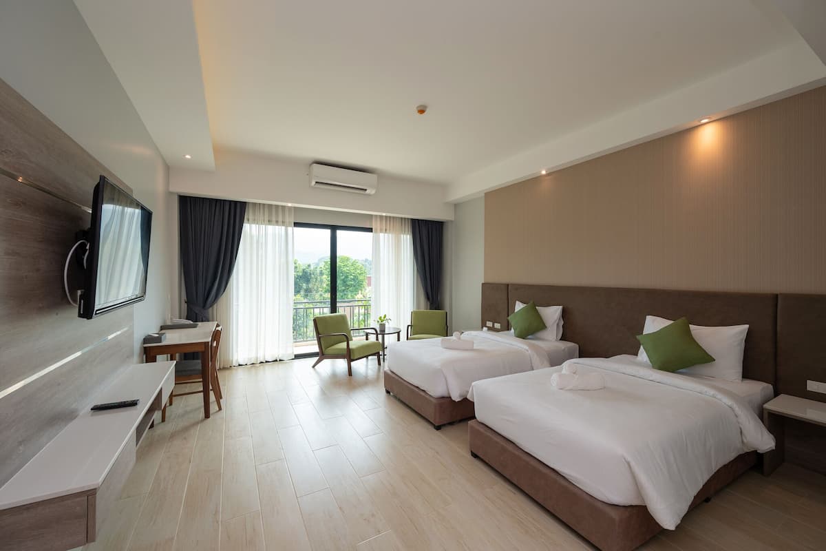 Habitación de hotel con dos camas, un escritorio, sillas, televisor y balcón con cortinas.