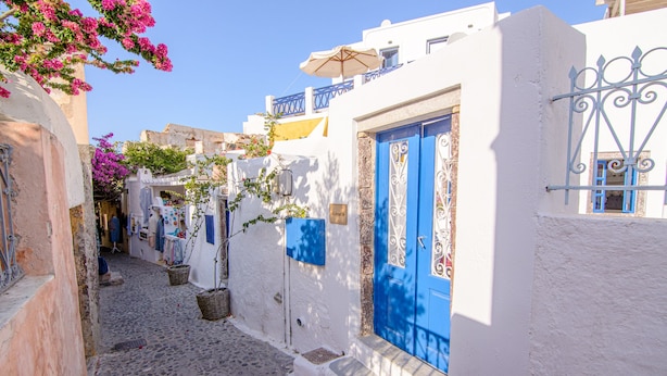 A narrow cobblestone street lined with white buildings, blue doors, and balconies with umbrellas. - ArcadOia (Santorini)