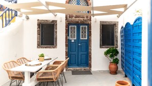 A small outdoor dining area with a white table and wicker chairs, a blue door, and a potted plant. - ArcadOia (Santorini)