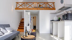 A modern living room with a sofa, a dining table with a bottle and glasses, a kitchen with a microwave and a stove, and a wooden loft above the living area. - ArcadOia (Santorini)