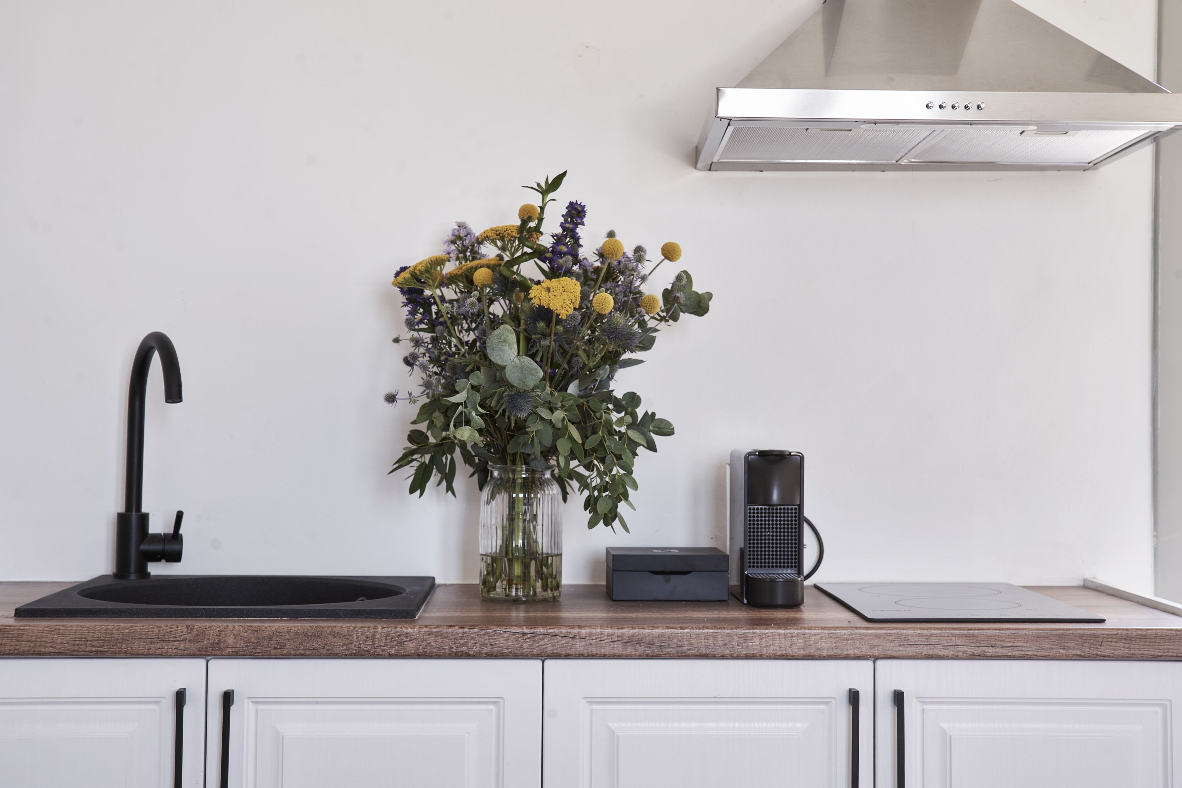 Een keuken met een zwarte kraan, een vaas met bloemen, een koffiezetapparaat en een afzuigkap.