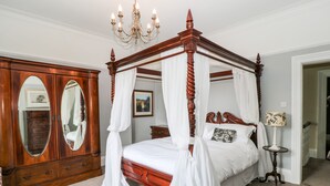 Uma cama de dossel com estrutura de madeira e roupa de cama branca, um armário espelho grande e um candelabro.