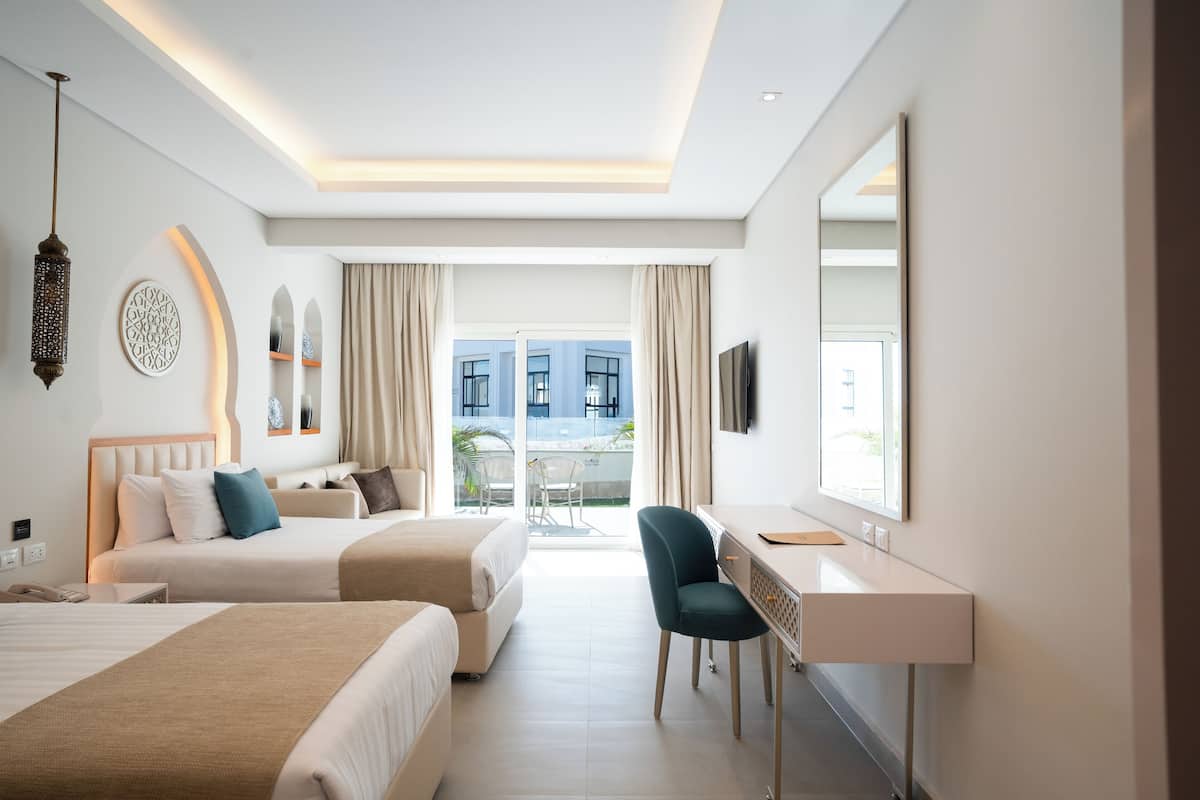Ein Hotelzimmer mit zwei Betten, einem Schreibtisch, einem Sessel, einem Fernseher und einem Balkon mit Blick auf Gebäude.