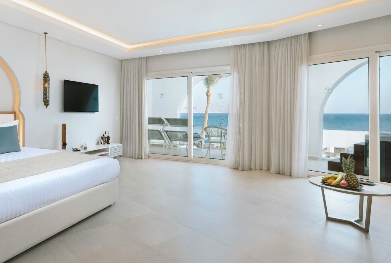 A modern hotel room with a large bed, a TV, a balcony with a view of the sea, and a small table with fruit.