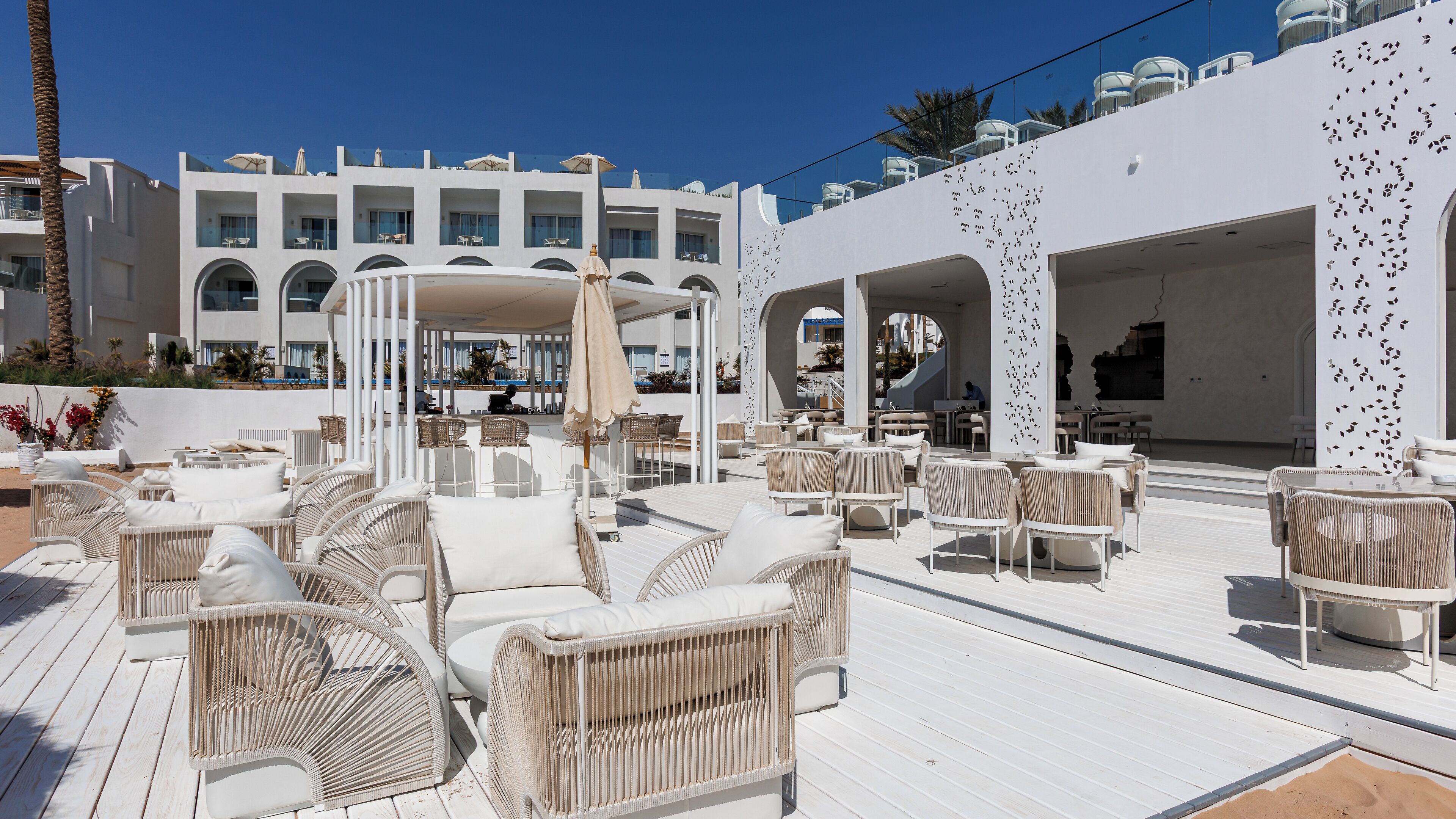 Um bar no terraço com móveis de vime e guarda-sóis, com vista para um edifício moderno com varandas.