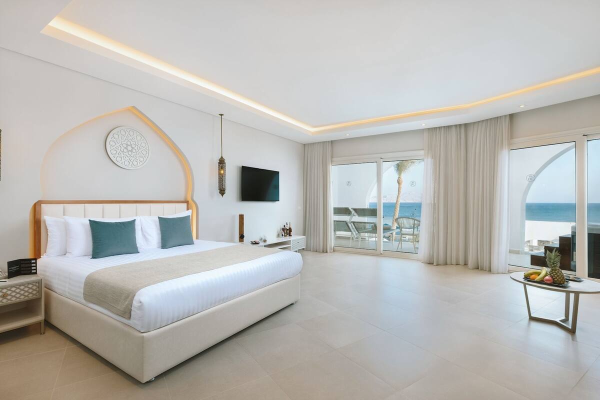 Ein modernes Hotelzimmer mit einem großen Bett, einem Fernseher, einem Balkon mit Meerblick und einem kleinen Tisch mit Obst.