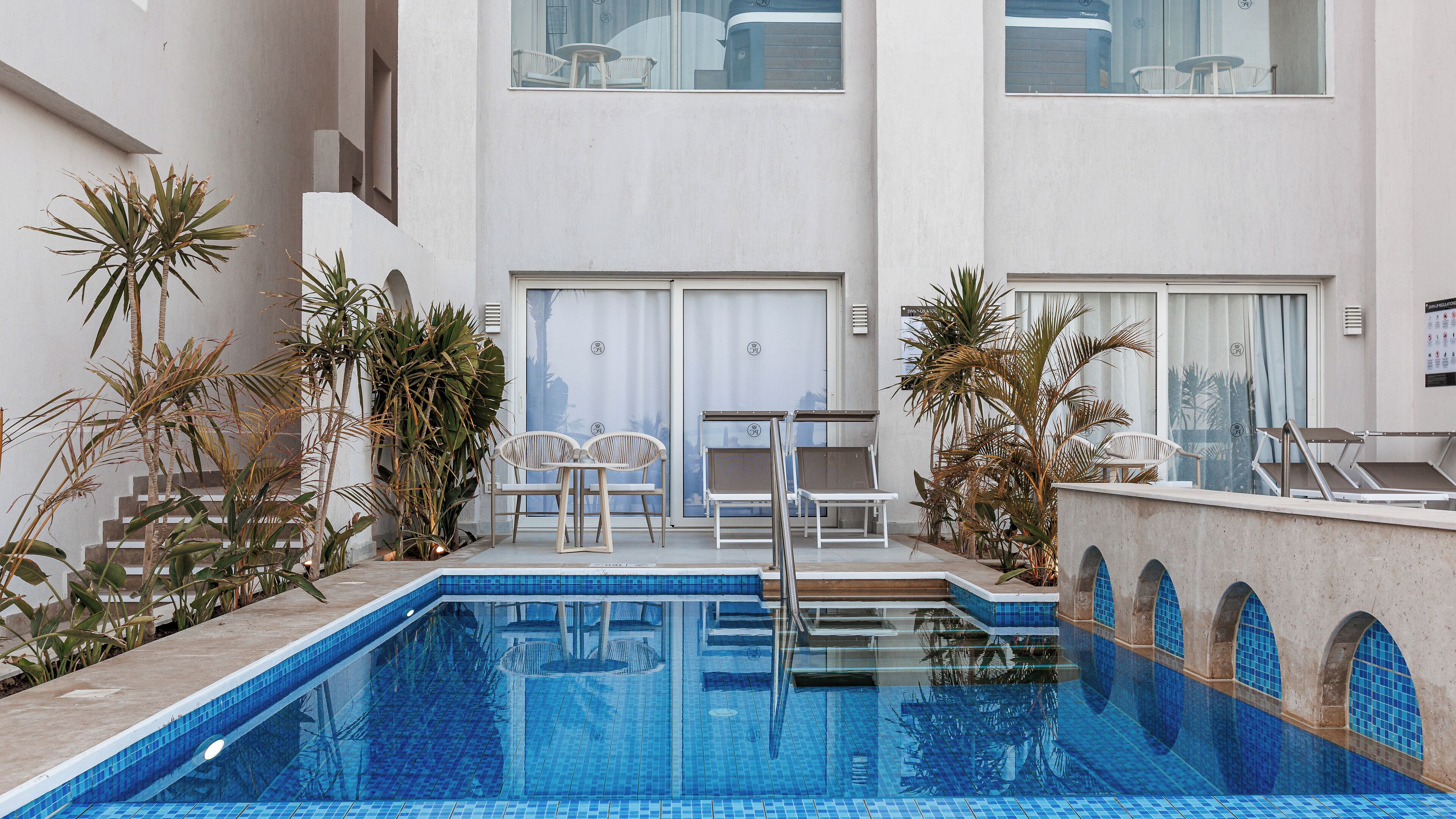 A swimming pool with steps leading into it, surrounded by potted plants and lounge chairs.