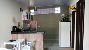 Une cuisine compacte avec un îlot central, des chaises blanches, une petite table, un réfrigérateur et une fenêtre avec des stores.