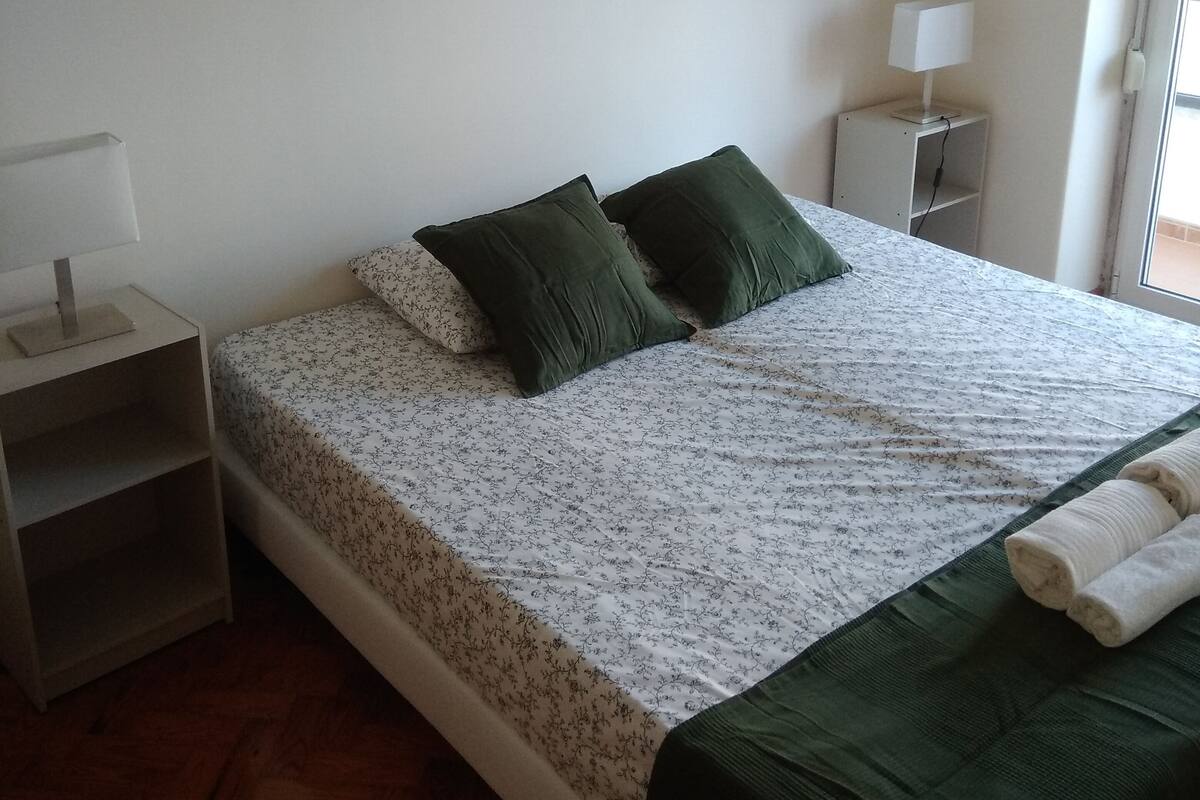 A bedroom with a bed, green pillows, a nightstand with a lamp, and two wall-mounted artworks.