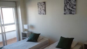 A room with two single beds, each with a green pillow and a white towel. There is a black hanging lamp, a small white table, and two wall art pieces. - Oriente DNA Studios V (Loures)