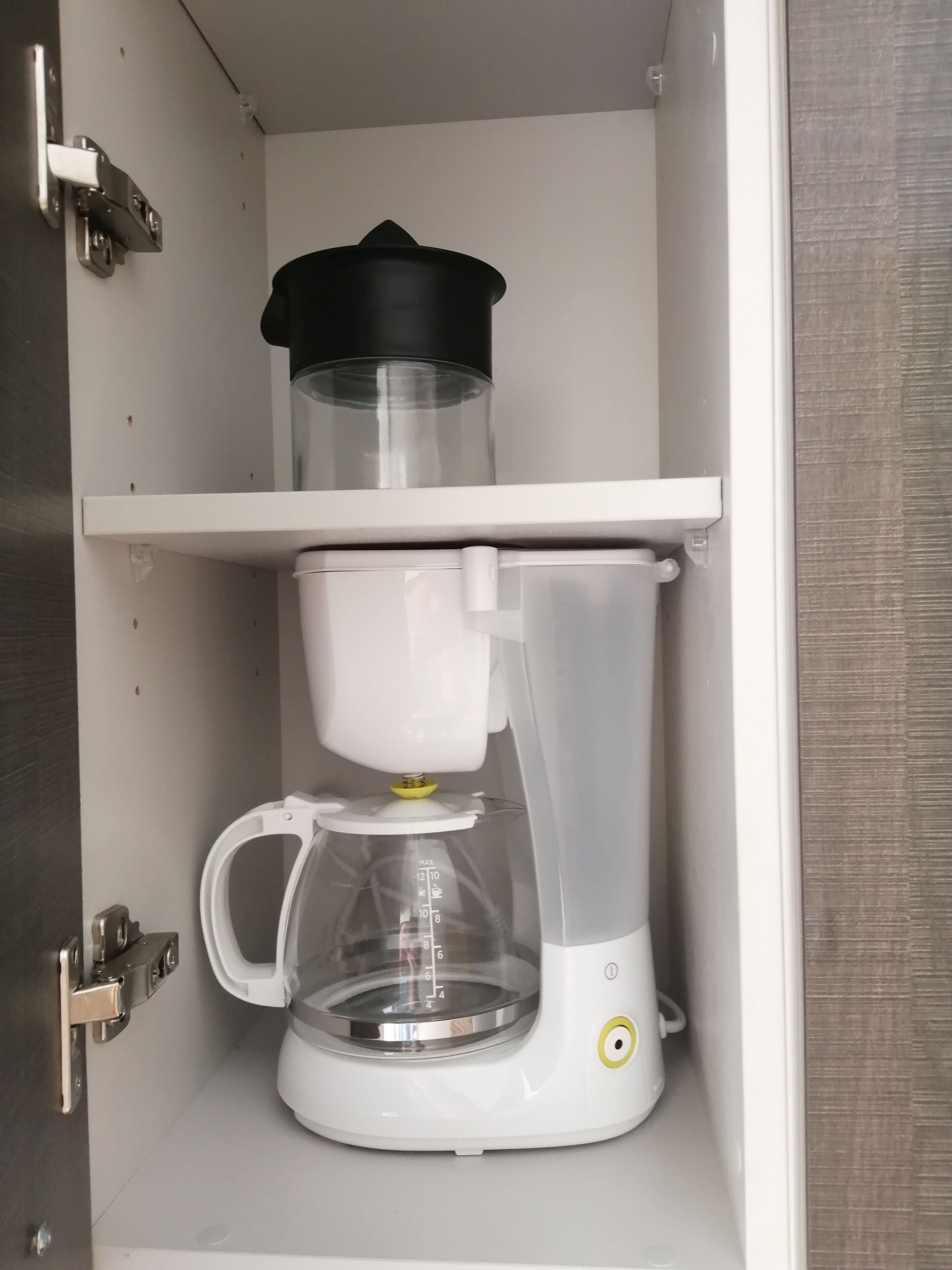A coffee maker with a transparent carafe and a black lid on top, placed on a white shelf in a cabinet.