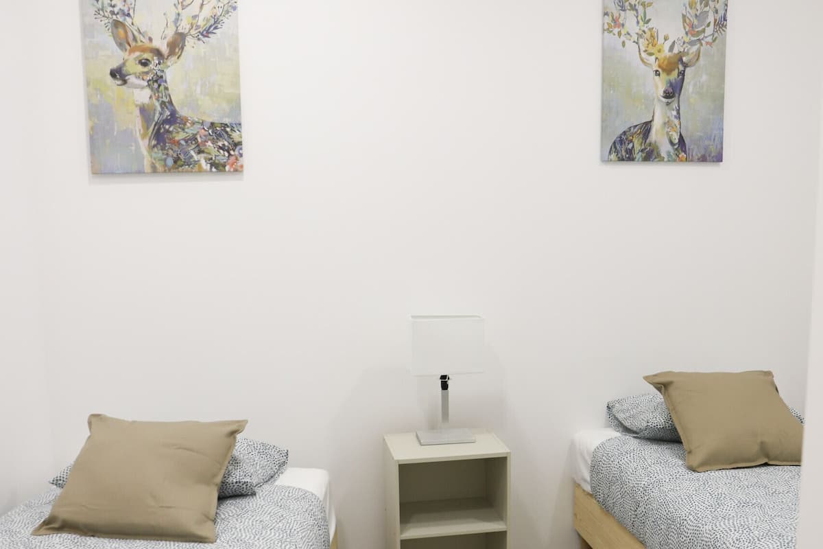 A bedroom with two beds, a bedside table with a lamp, and two wall-mounted paintings of deer.