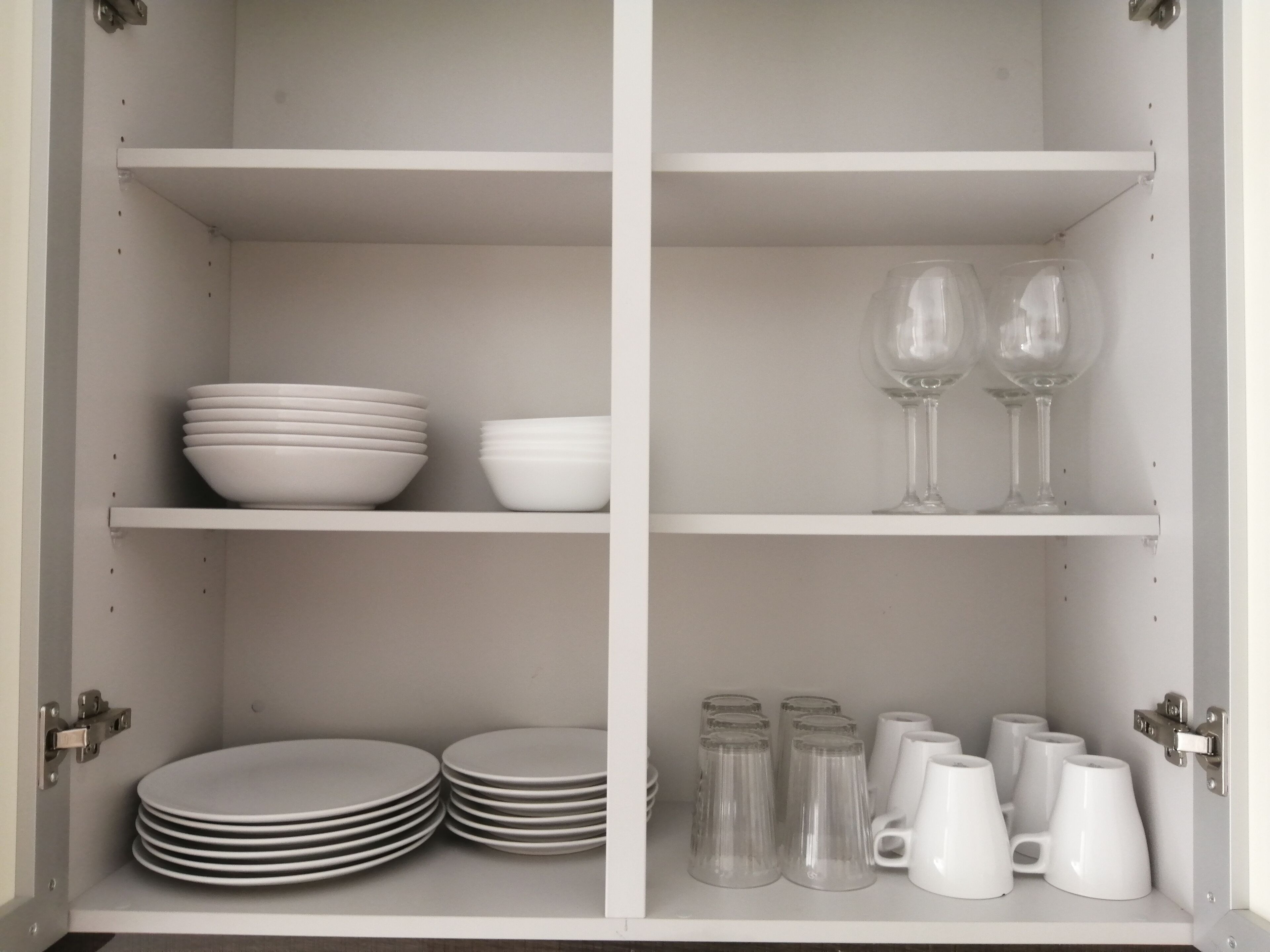 A white cabinet with shelves holding white plates, bowls, glasses, and mugs.