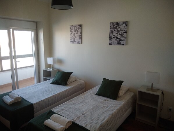 A room with two single beds, each with a green pillow and a rolled white towel. There is a small bedside table with a lamp, and two wall-mounted artworks.
