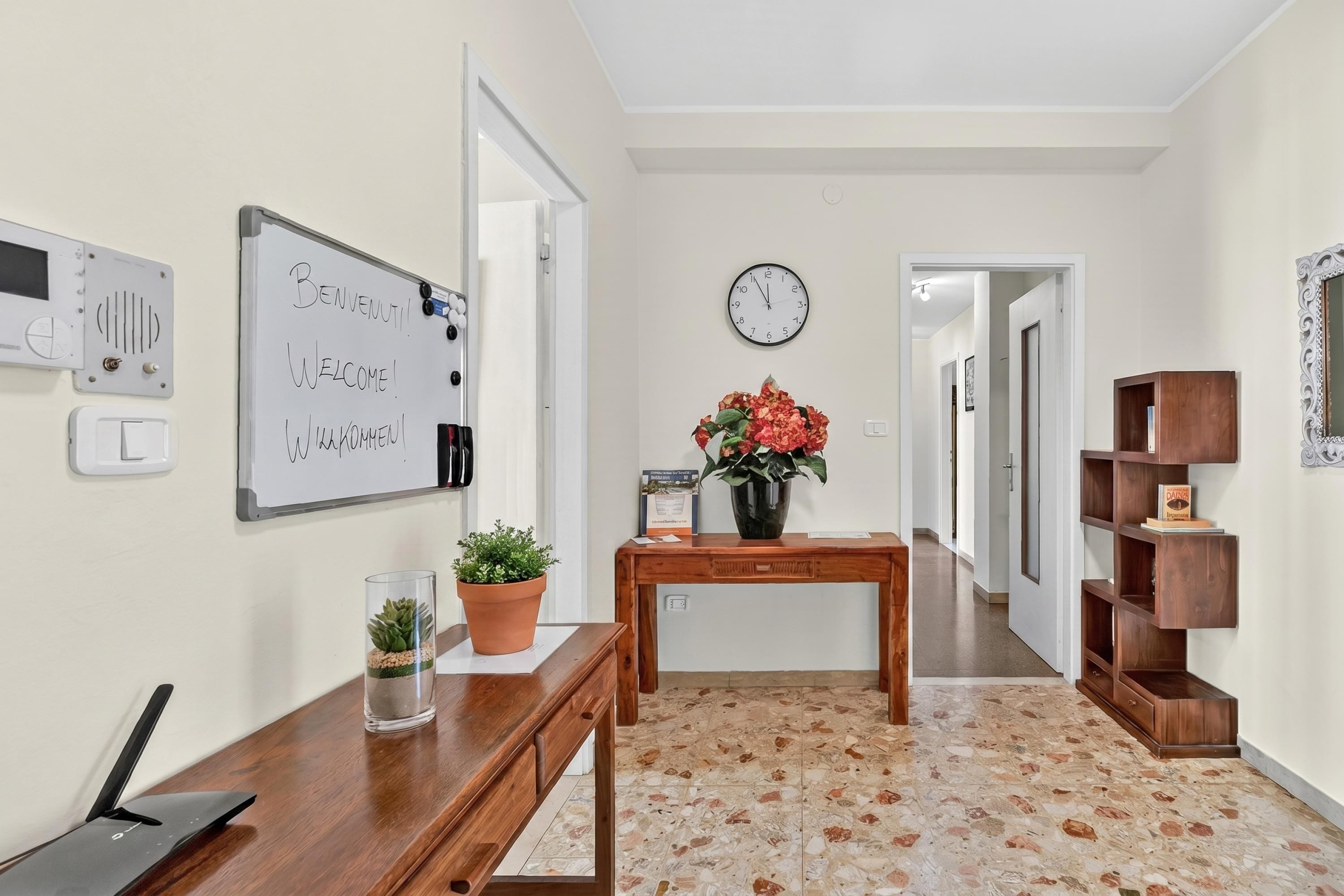 A hallway with a whiteboard, a clock, a wooden table, a potted plant, and a shelf unit.
