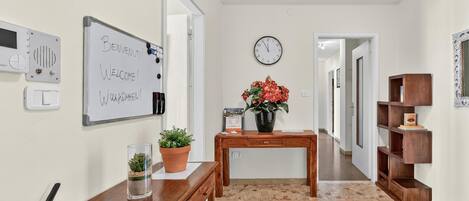 A hallway with a whiteboard, a clock, a wooden table, a potted plant, and a shelf unit.