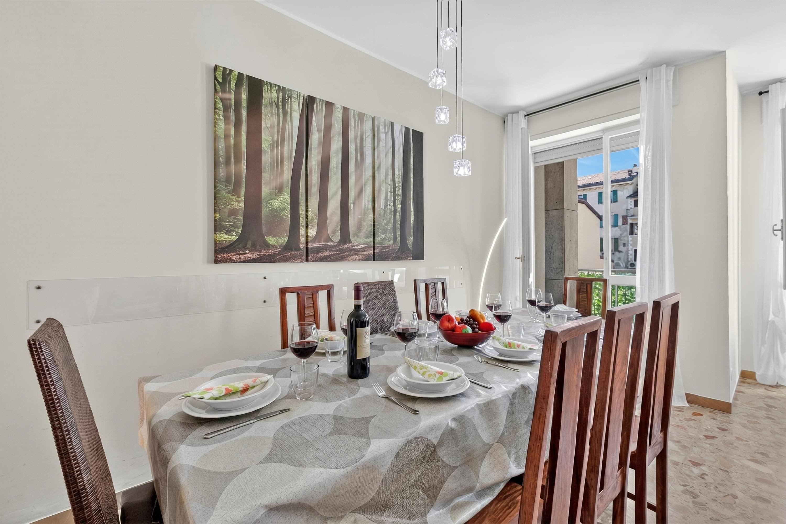 A dining area with a table set for six, a painting of a forest on the wall, and a view of buildings outside.
