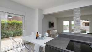 Une cuisine moderne dotée d’un grand îlot central, d’un coin repas avec des chaises transparentes et d’un téléviseur fixé au mur.