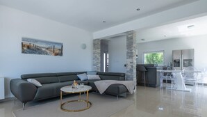 Un salon moderne avec un canapé d’angle gris, une table basse ronde et un coin repas composé de chaises transparentes et d’une table blanche.