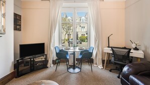 Een woonkamer met een televisie, een eettafel met stoelen, een bureau met stoel en uitzicht op gebouwen door een raam.
