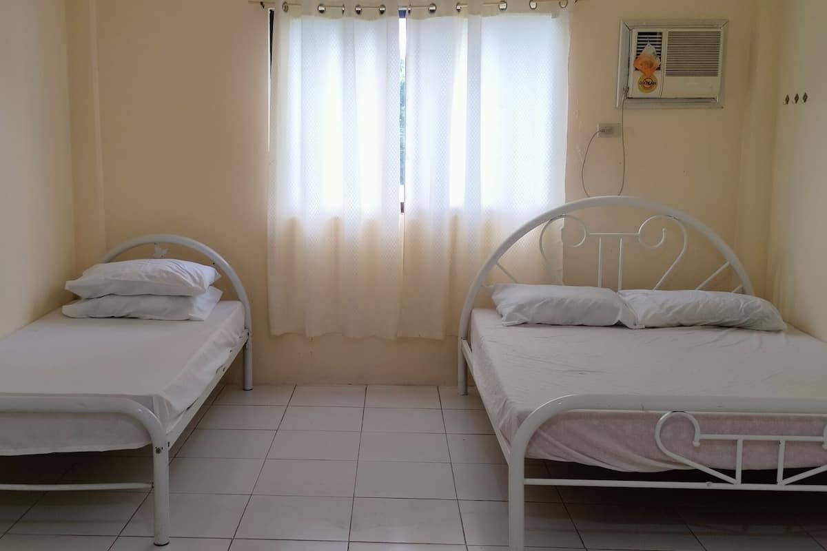 Ein Zimmer mit zwei Einzelbetten, einem mit Vorhängen versehenen Fenster, einer Wand-Klimaanlage und einem gefliesten Boden.