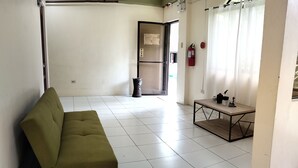 Ein Zimmer mit einem grünen Sofa, einem kleinen Tisch mit Dekoration, einem Feuerlöscher und einer Tür zu einem anderen Raum.