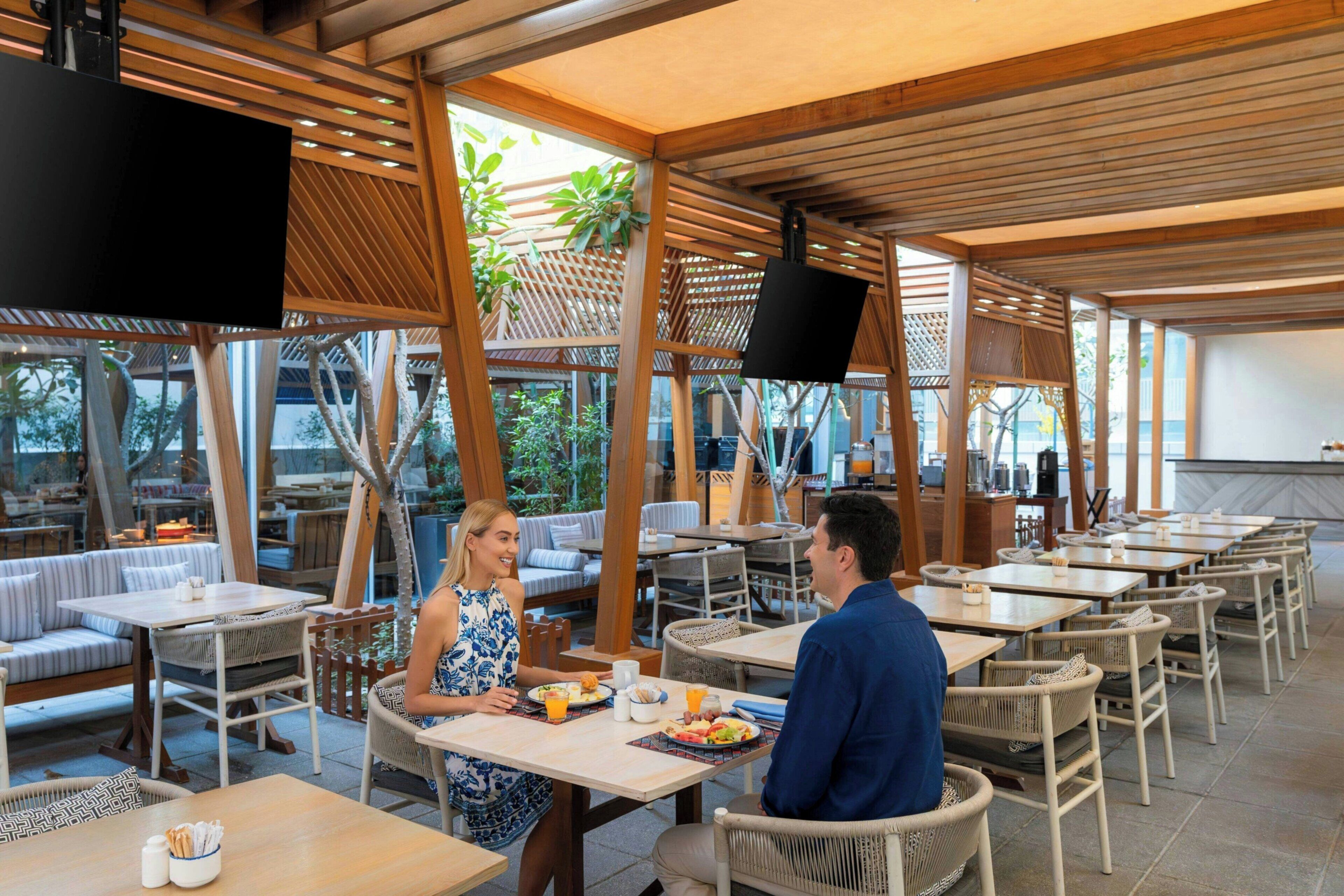 An outdoor dining area with wooden tables and chairs, a TV screen, and a view of the outdoors.