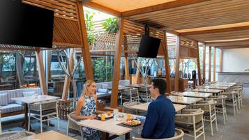 An outdoor dining area with wooden tables and chairs, a TV screen, and a view of the outdoors.