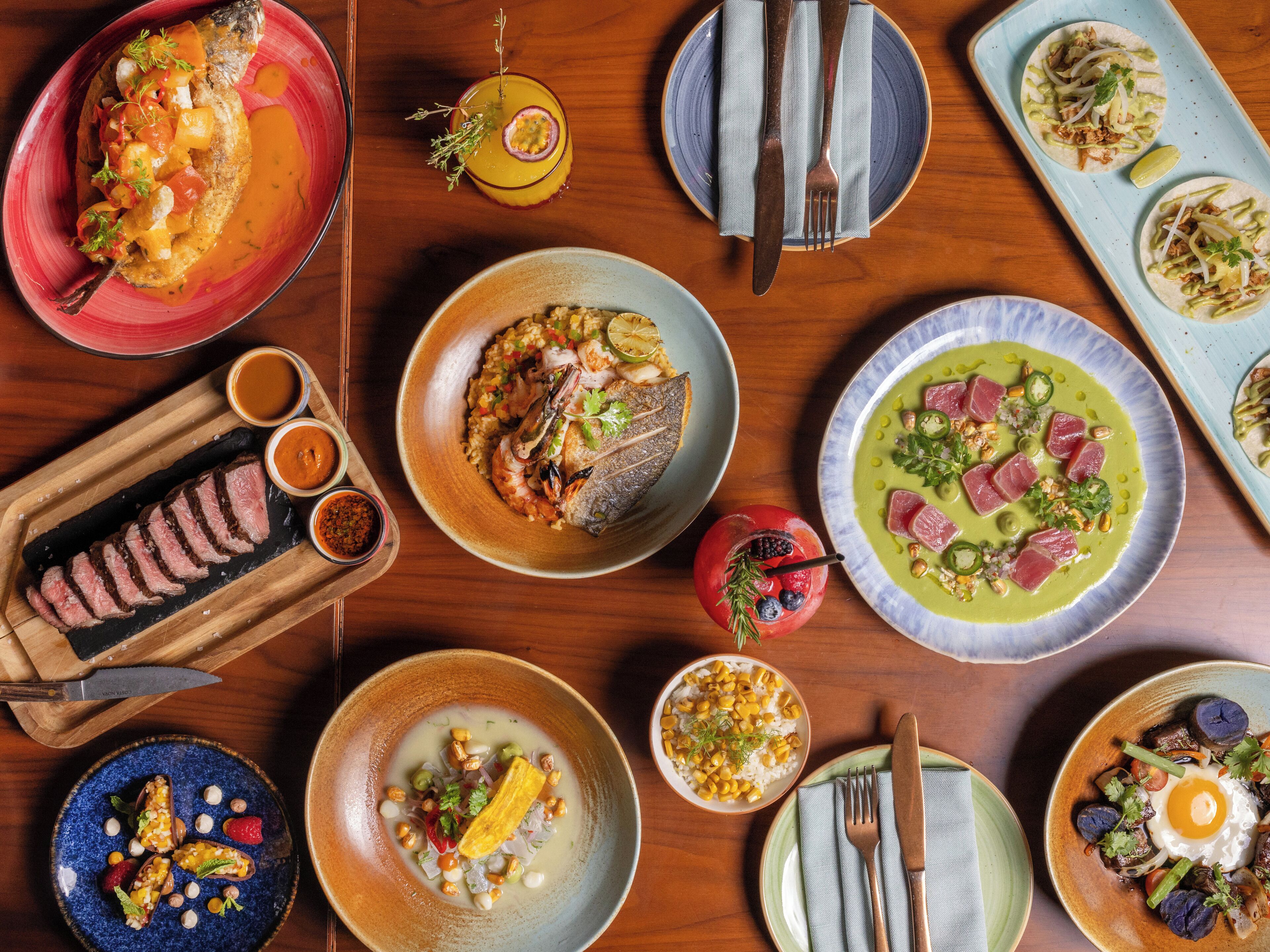 Diversos pratos sobre uma mesa de madeira, incluindo peixe, tacos e uma tigela de milho.
