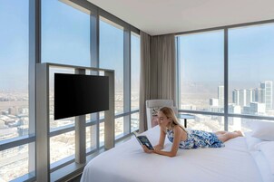 Habitación de hotel con una cama grande, un televisor de pantalla plana montado en la pared y vistas a la ciudad a través de amplias ventanas.