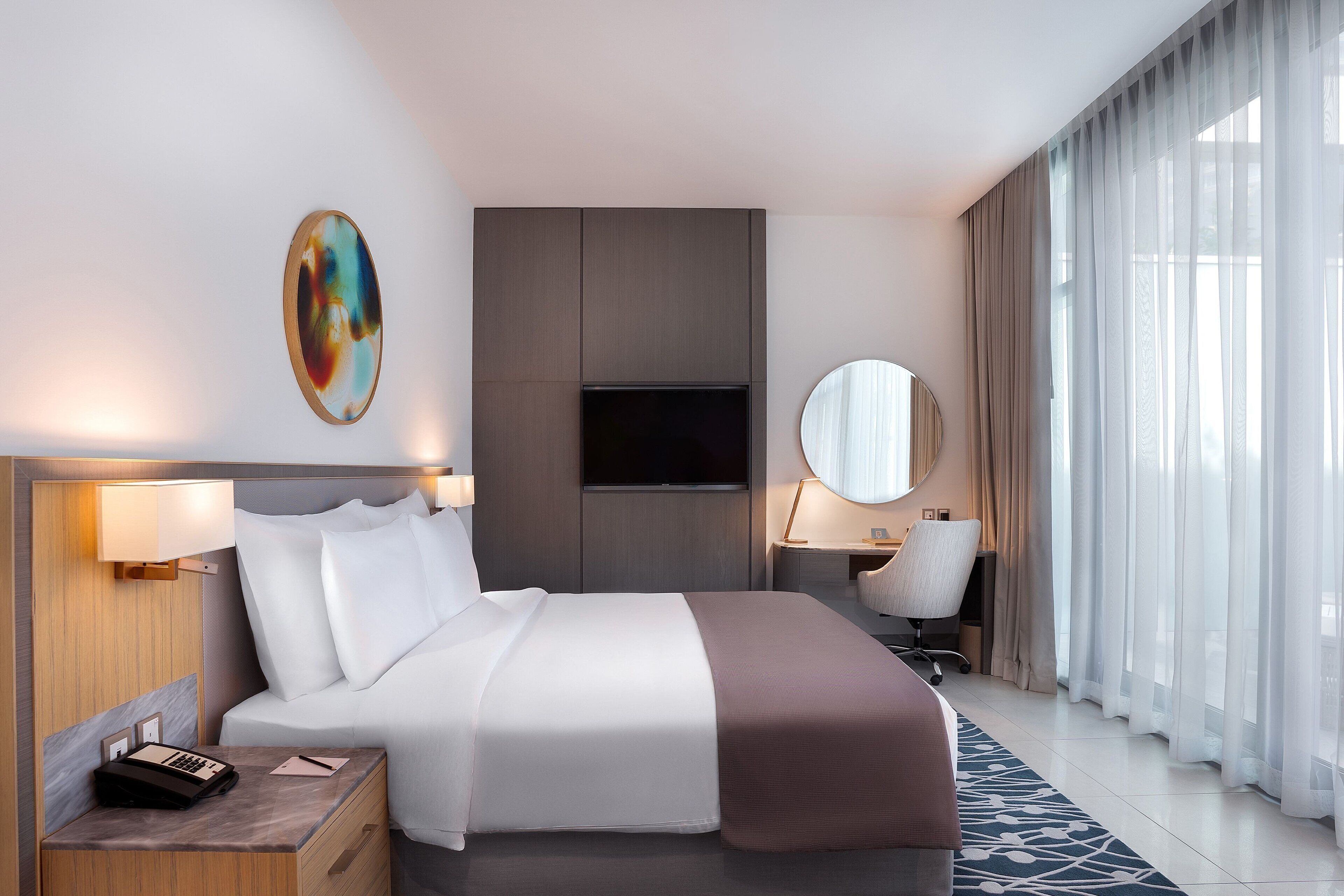 Una habitación de hotel moderna con una cama grande, un escritorio con lámpara, una silla, un televisor y un ventanal con cortinas traslúcidas.
