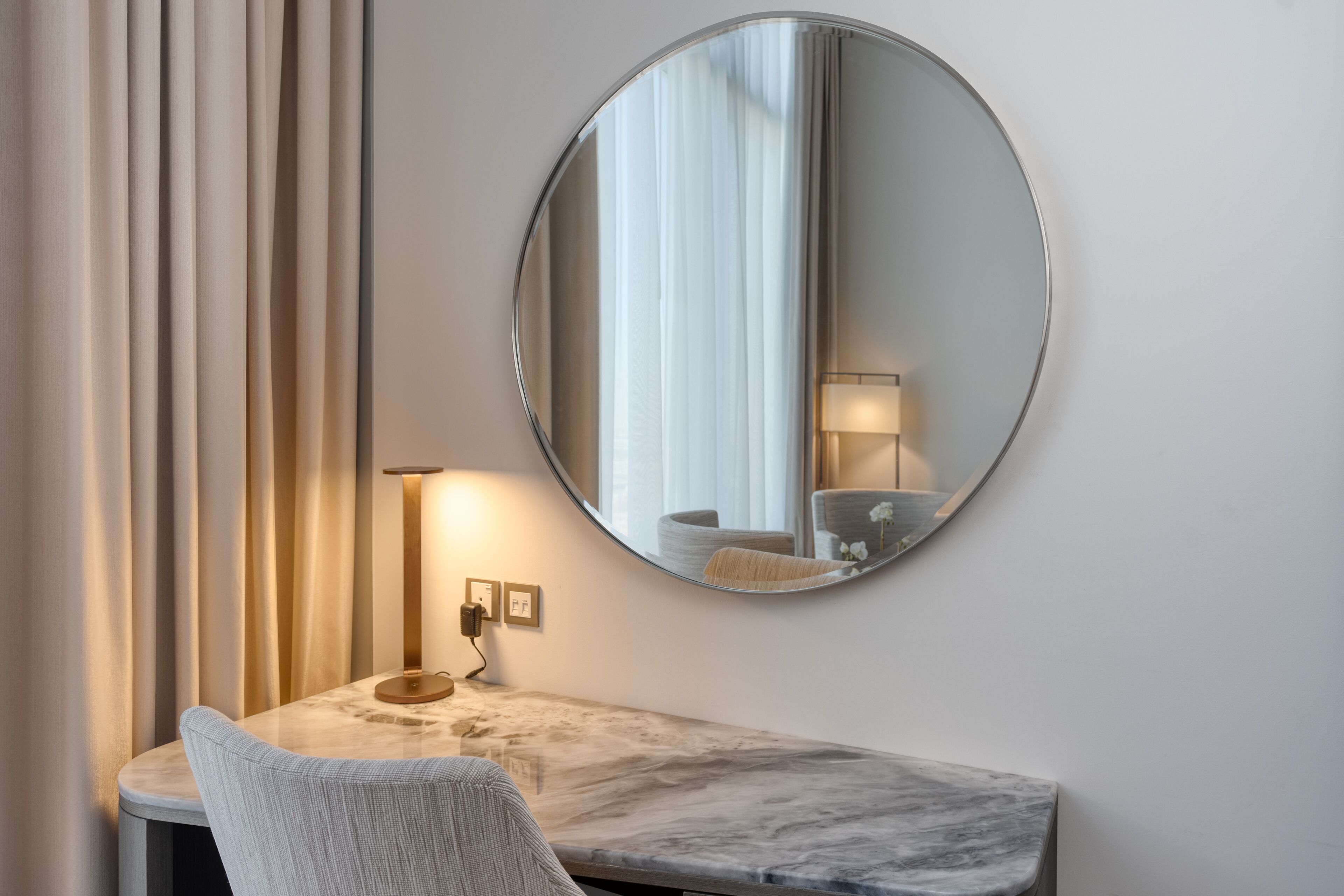 A modern room with a marble desk, a round mirror, a lamp, and a chair.