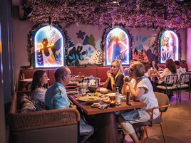 Un restaurante con cabinas y mesas, con un techo decorado con follaje colgante y obras de arte en las paredes.