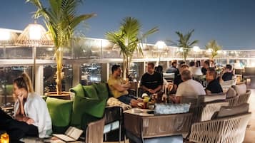 A rooftop bar with outdoor seating, palm trees, and city views at night.