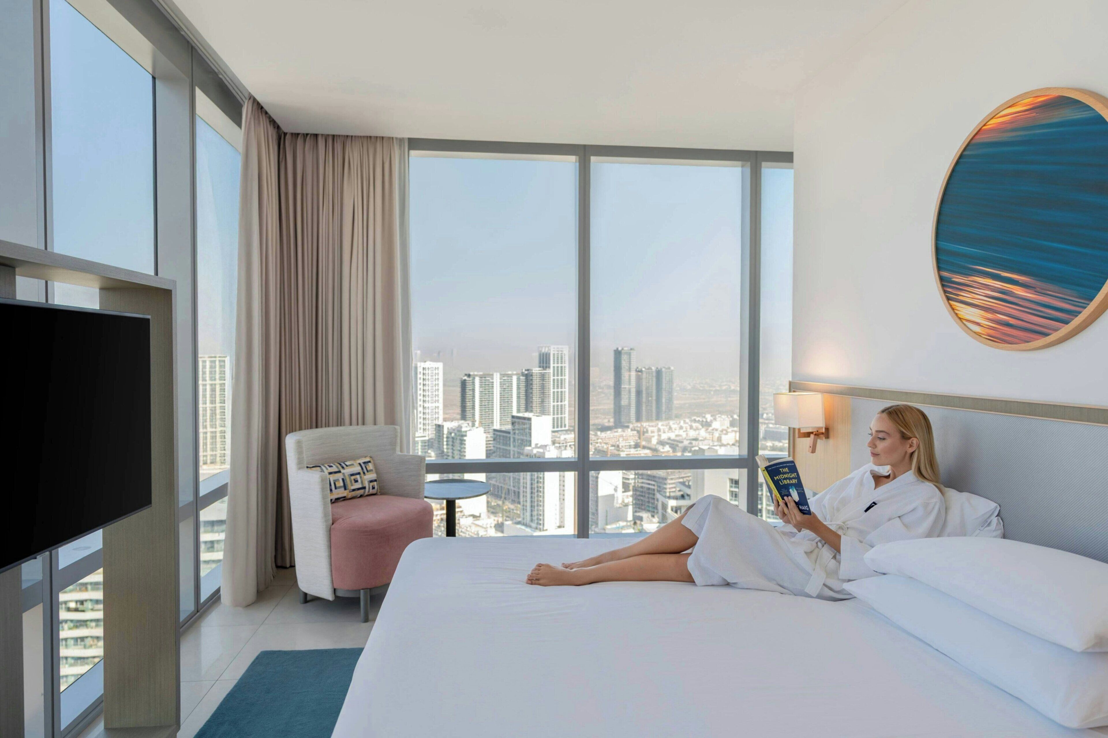 A hotel room with a large bed, a TV, a chair, a table, and a city view.