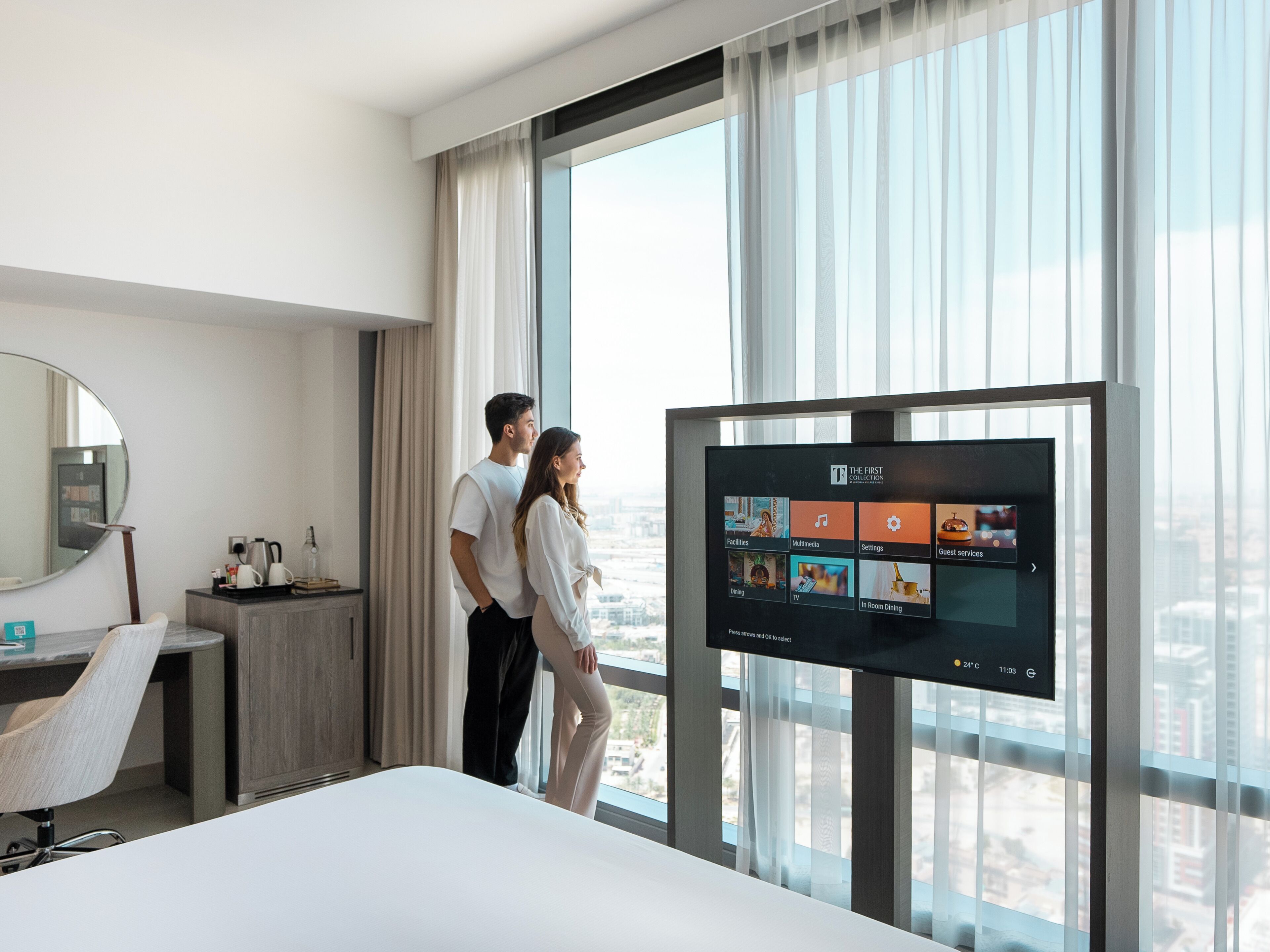 Ein Hotelzimmer mit großem Fenster, einem Bett, einem Fernseher, der Zimmerauswahl anzeigt, einem Schreibtisch mit Stuhl und einem Spiegel.