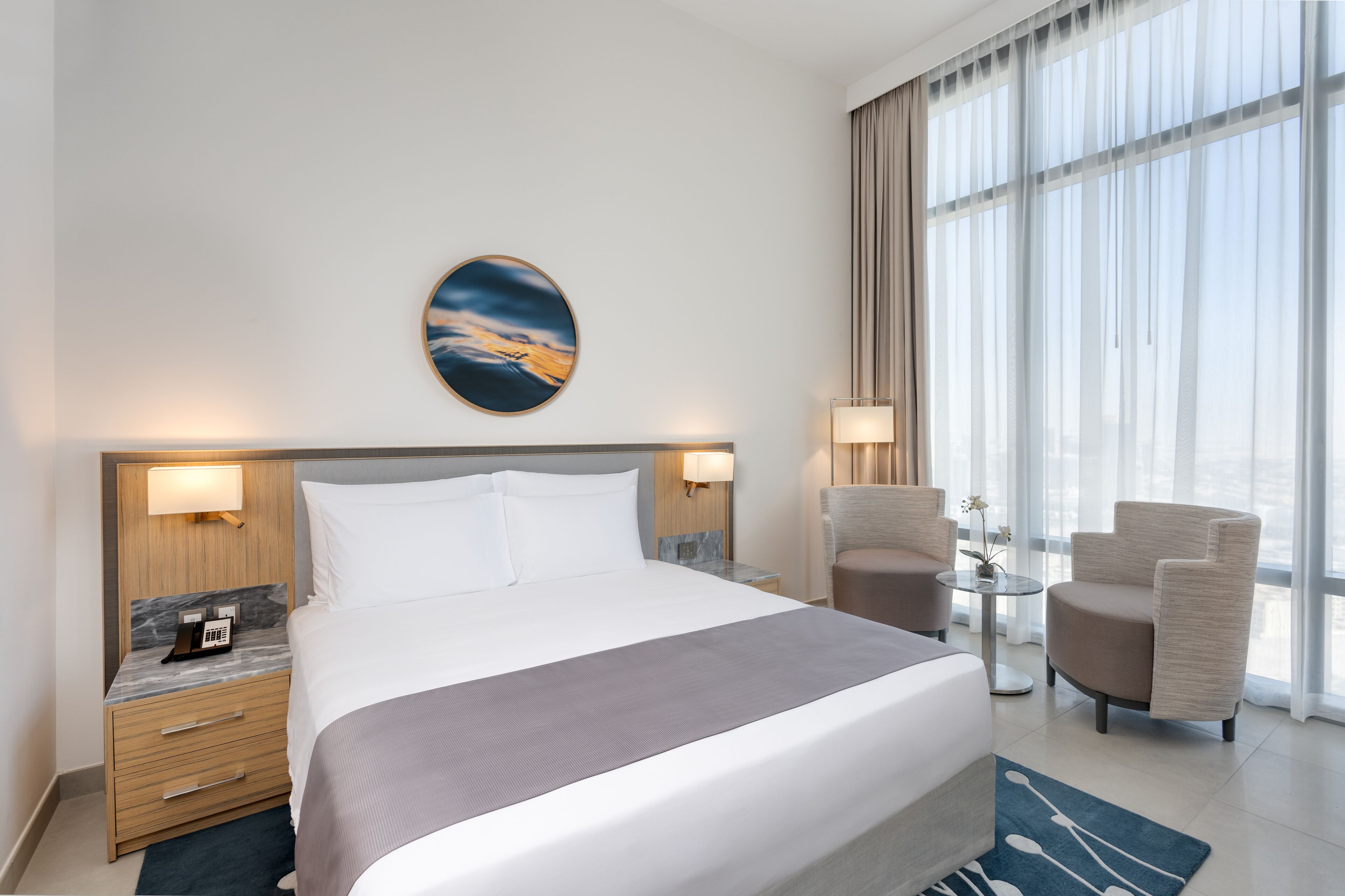 A modern hotel room with a large bed, a nightstand with a phone, a small table with a lamp, and a chair.