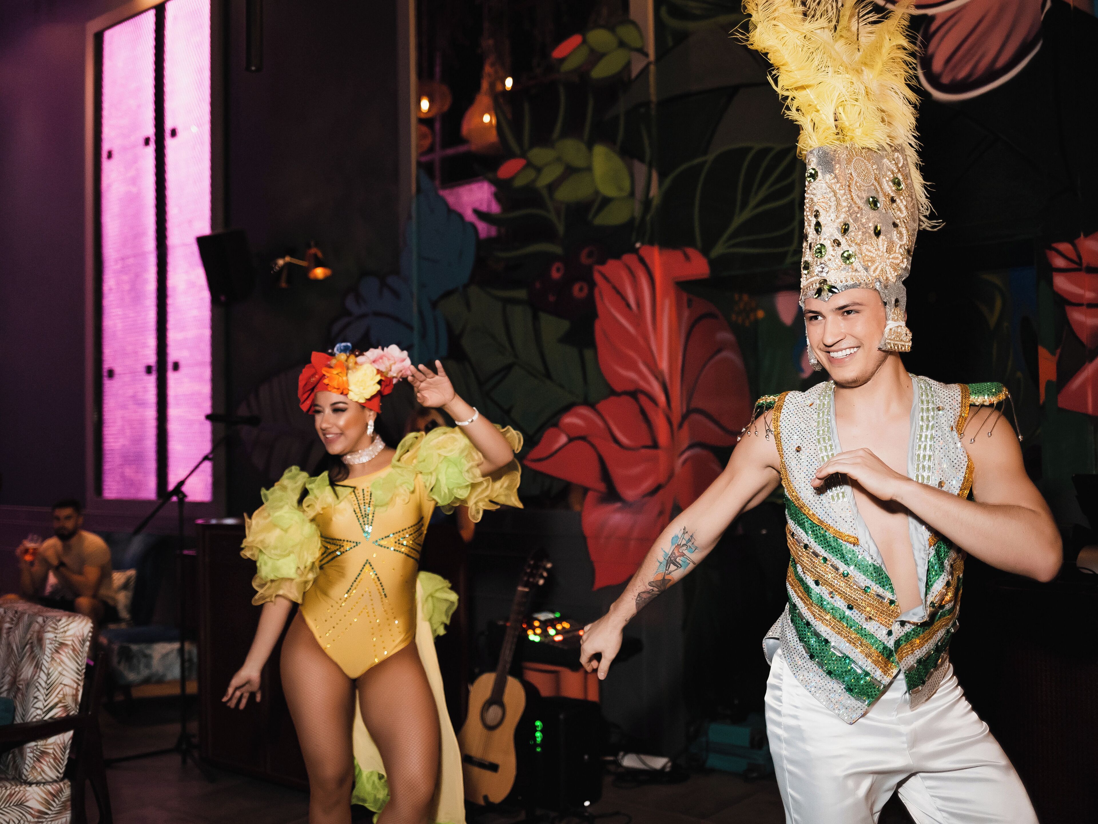 Two performers in colorful costumes with a mural in the background.