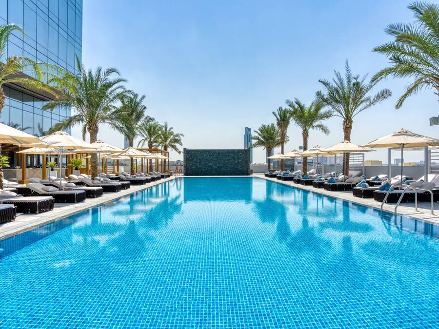 A swimming pool with lounge chairs and umbrellas, surrounded by palm trees and a modern building.