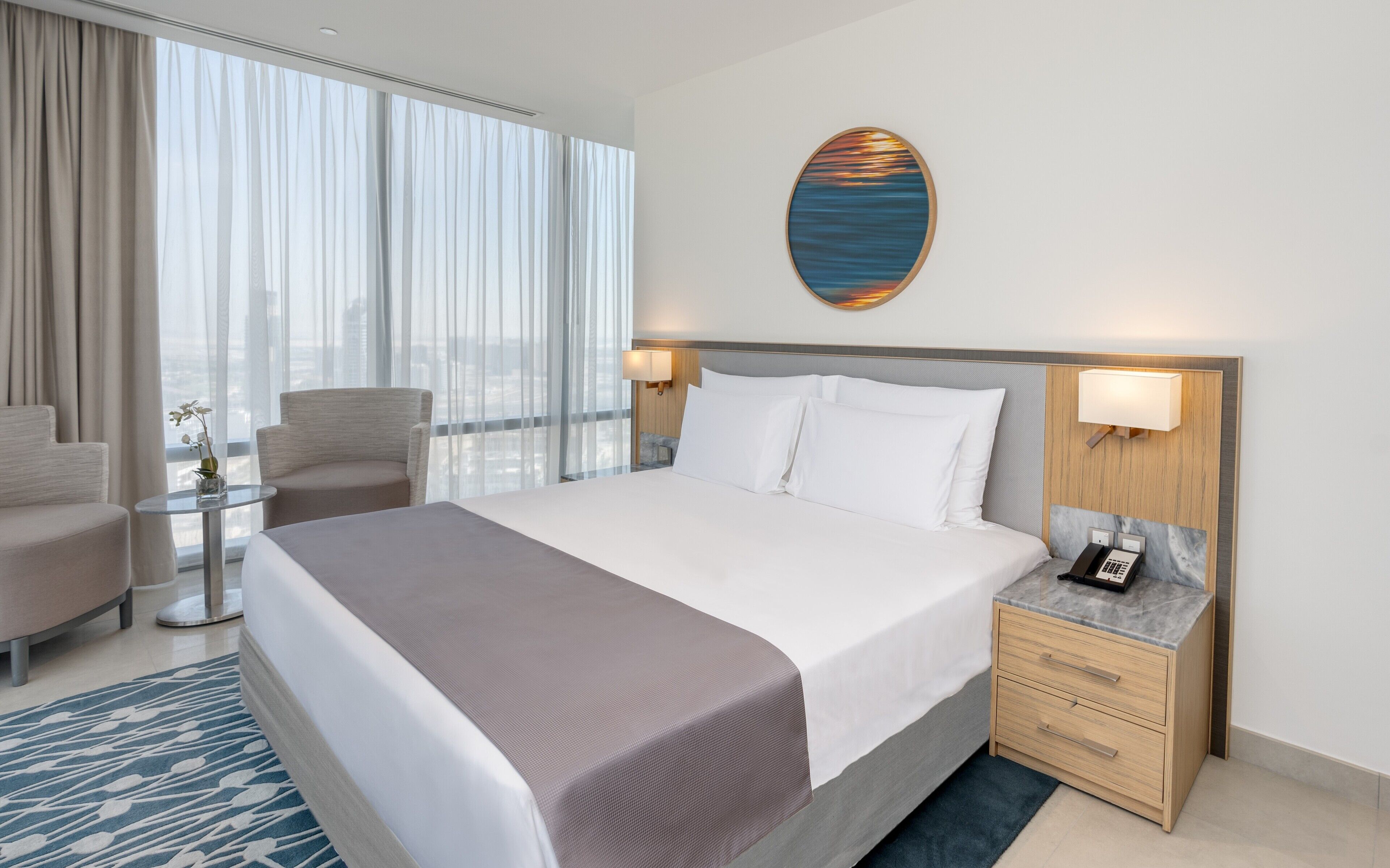 A hotel room with a large bed, a bedside table with a phone, a small table with a vase, and a chair.