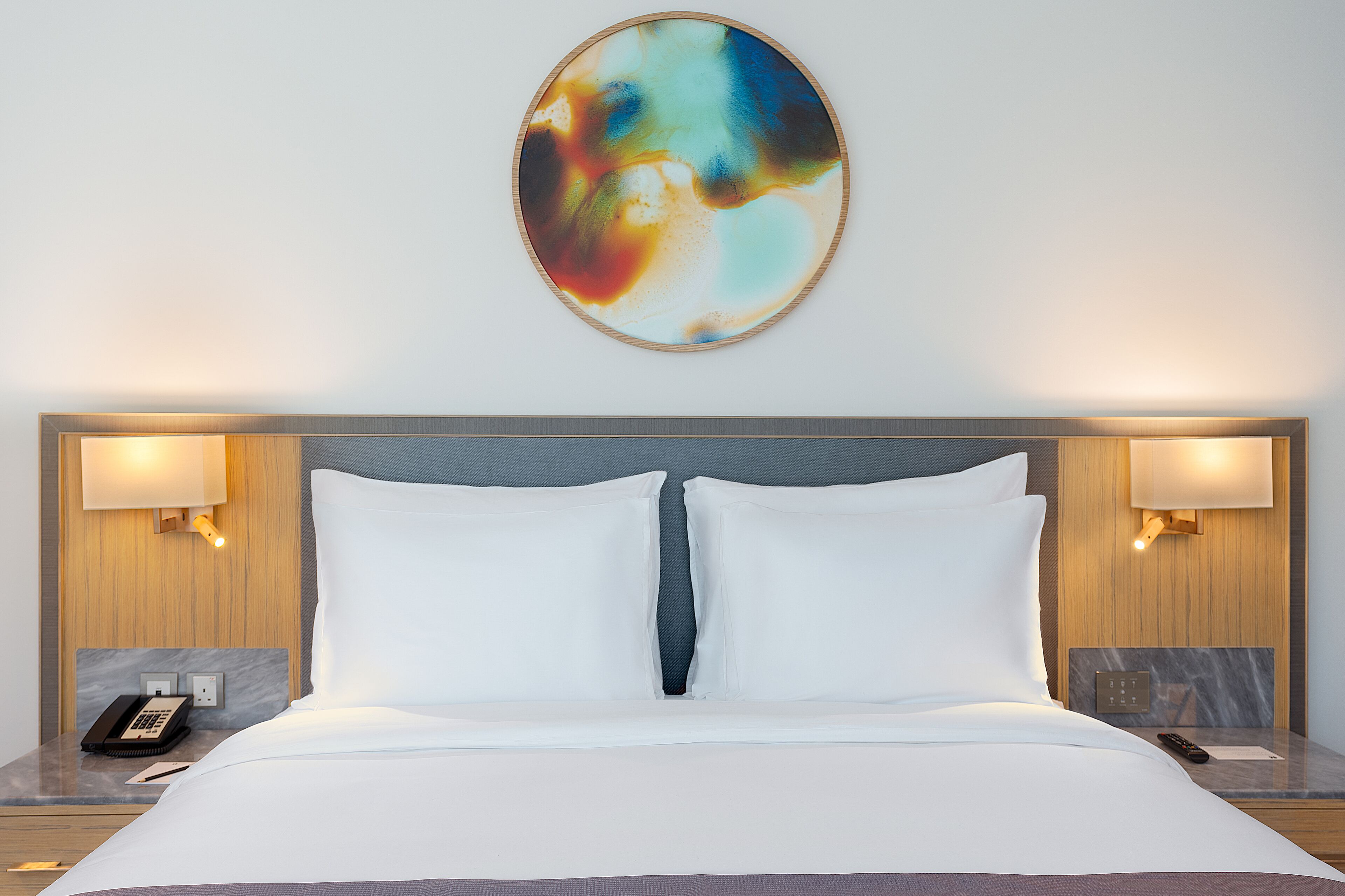 A hotel room with a large bed, two bedside lamps, a telephone, and a wall-mounted abstract painting.