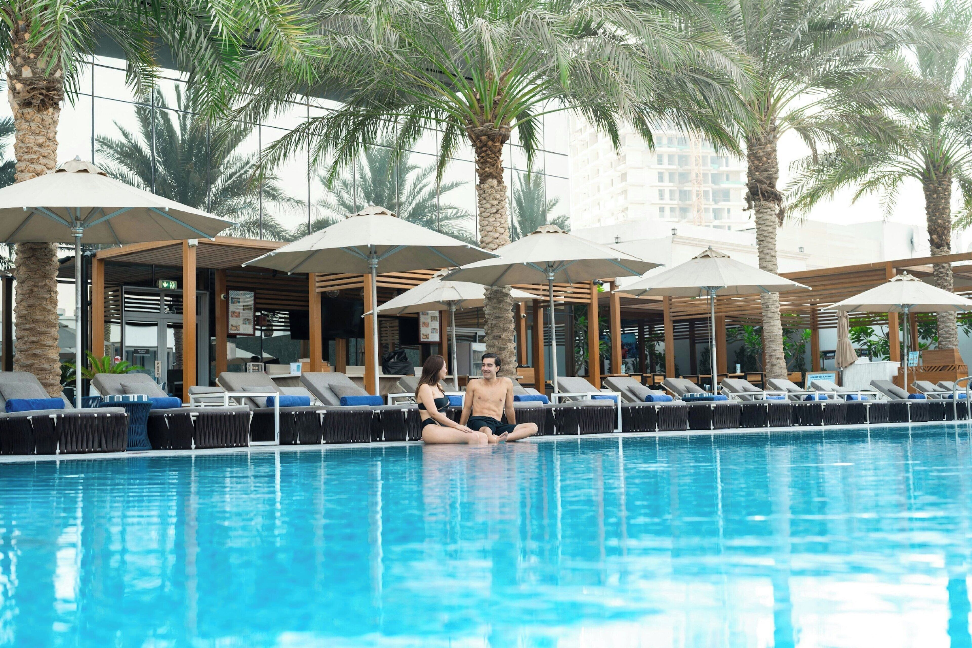 A pool area with lounge chairs and umbrellas, surrounded by palm trees and a modern building in the background.
