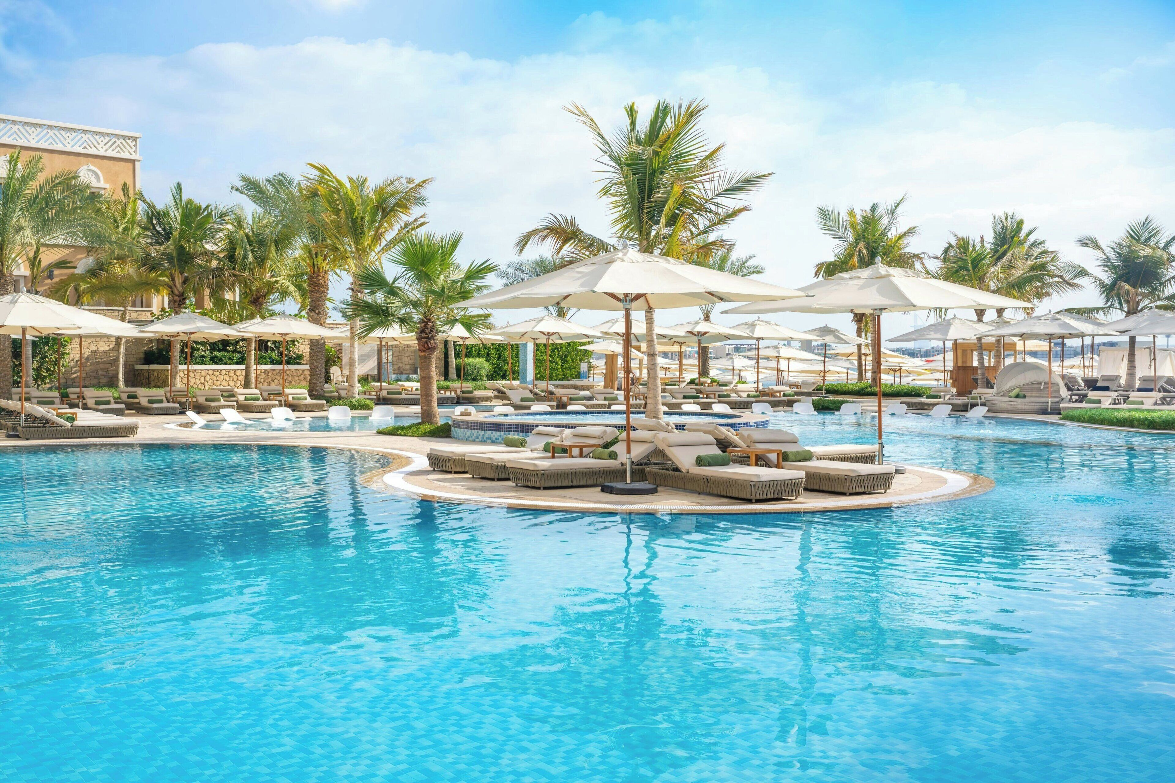 A resort pool area with multiple lounge chairs and umbrellas, surrounded by palm trees.