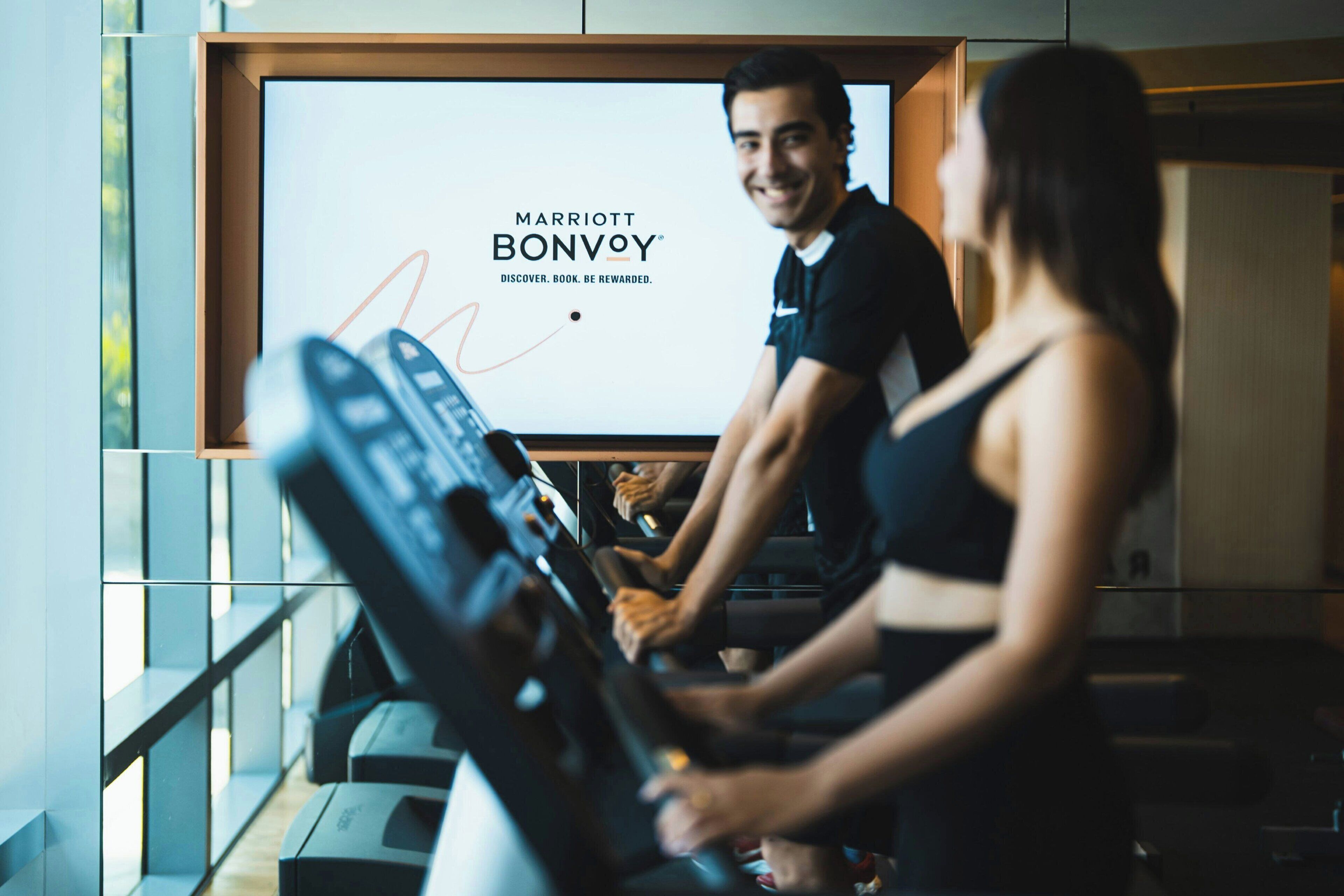 Un gimnasio con una pantalla grande que muestra el logotipo de Marriott Bonvoy y una caminadora en uso.