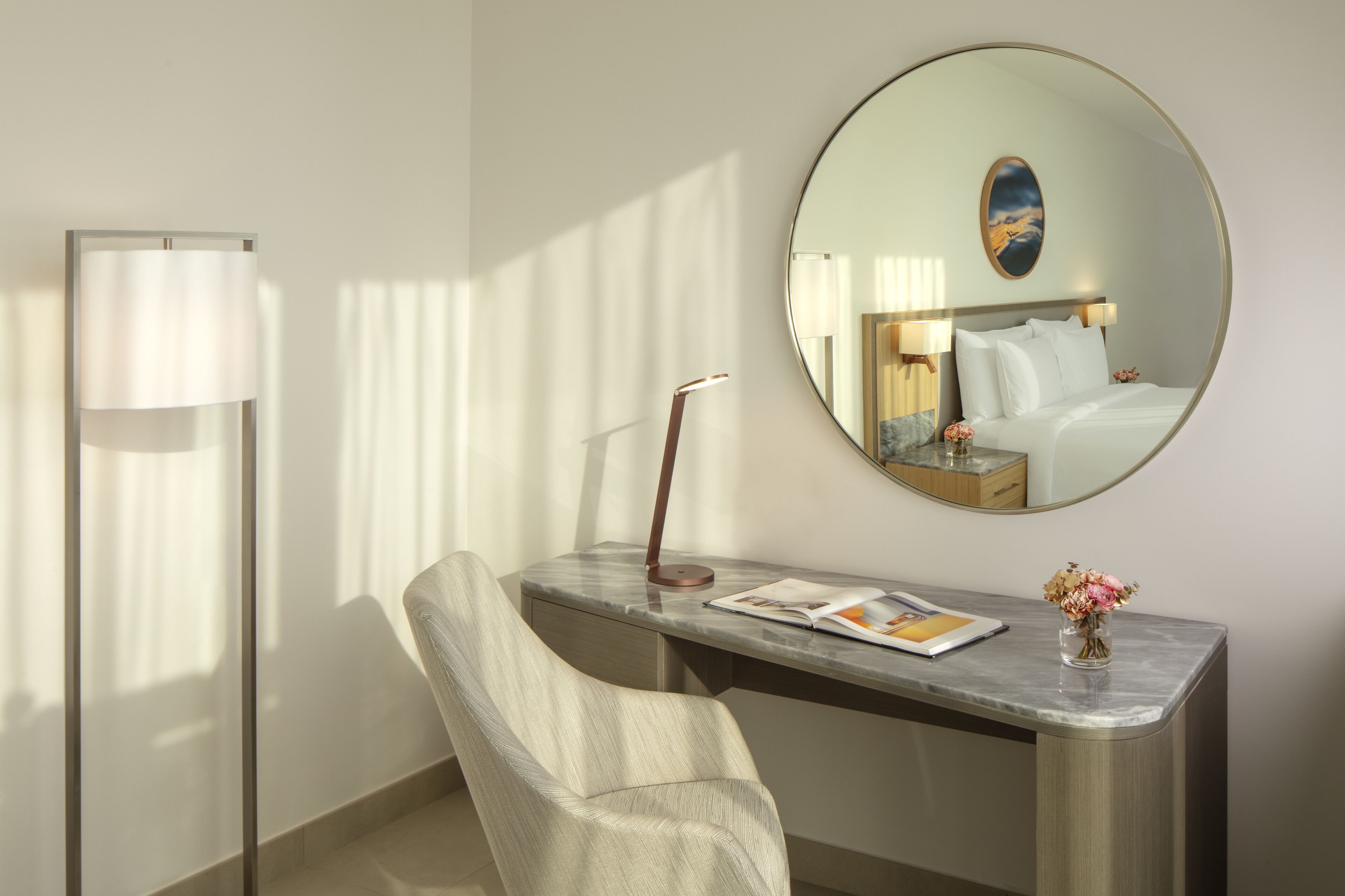 Une chambre d’hôtel avec un lit, un bureau doté d’une lampe, une chaise, un miroir rond et un vase avec des fleurs.