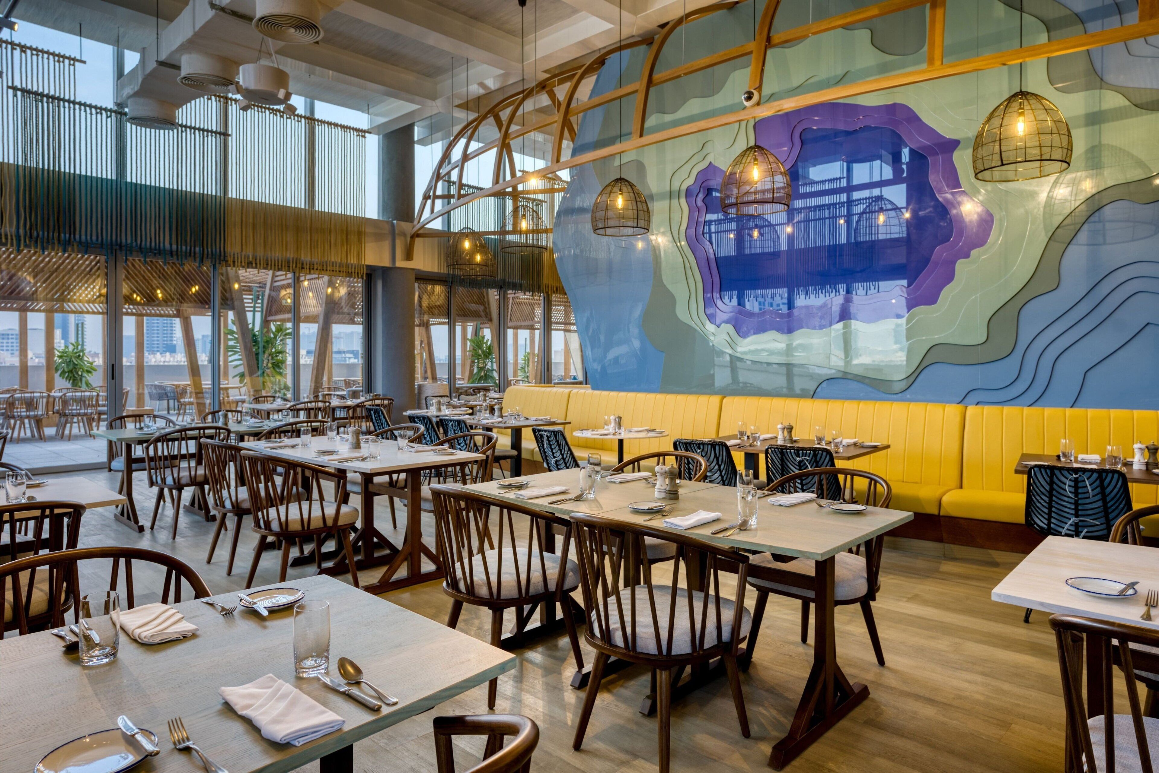 A restaurant with multiple tables and chairs, large windows, and a mural on the wall.